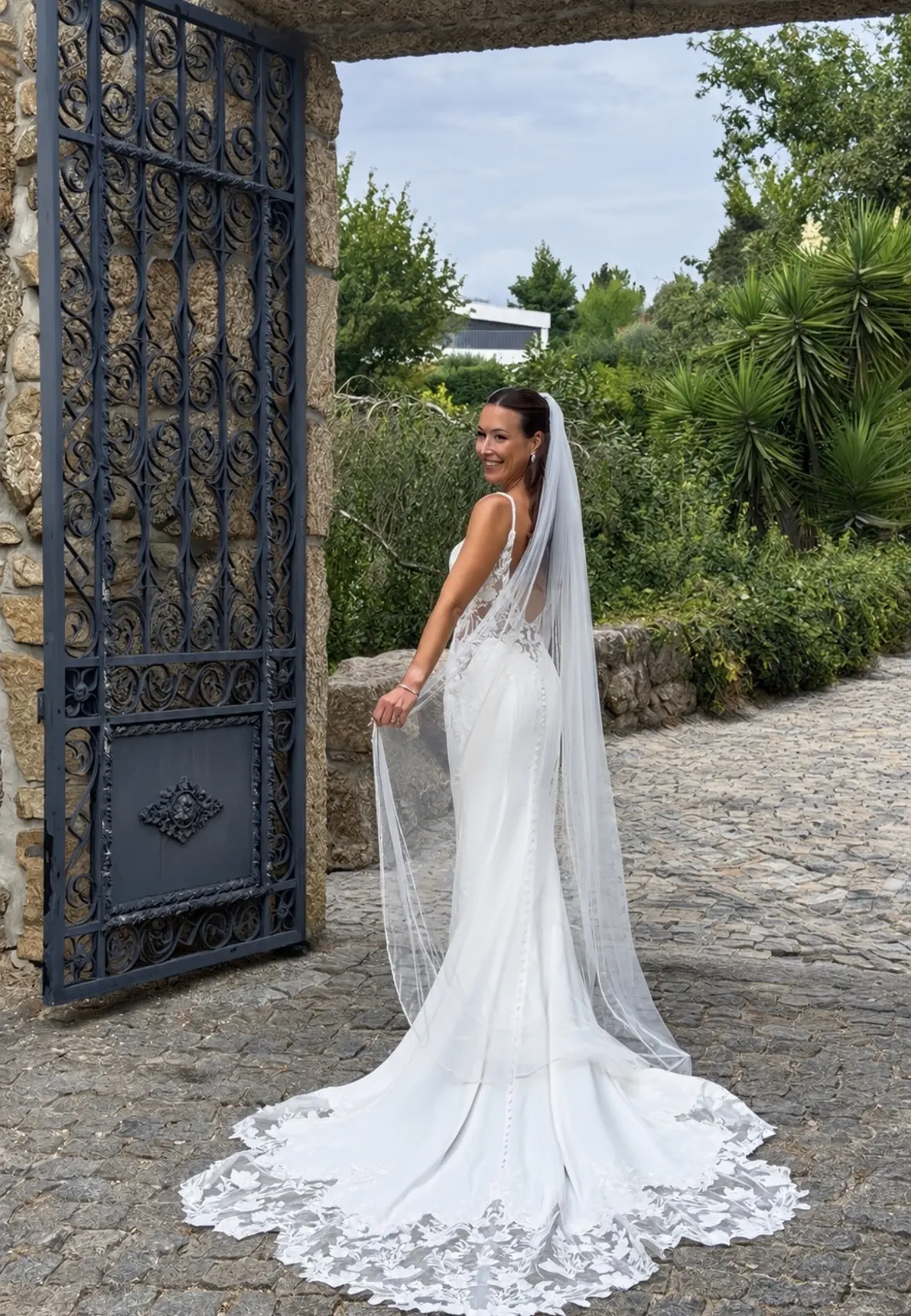 Mariée souriante de dos devant un portail en fer forgé, voile long et traîne en dentelle
